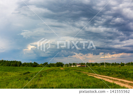 Dramatic stormy clouds loom over a lush green meadow, creating an intense sky above a quiet dirt road in the countryside during a summer day before the impending rain falls Dramatic stormy clouds loom over a lush green meadow, creating an intense sky above a quiet dirt road in the countryside during a summer day before the impending rain falls 141555562