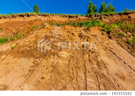 Low angle view of a weathered sandy quarry slope with textured soil erosion, sparse green grass, and vibrant trees against a clear blue summer sky, raw natural geological formation 141555582