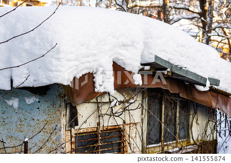 Thick white snow blankets the roof of a weathered, rustic shed, contrasting sharply with rusty metal walls and bare winter branches Thick white snow blankets the roof of a weathered, rustic shed, contrasting sharply with rusty metal walls and bare winter branches 141555874