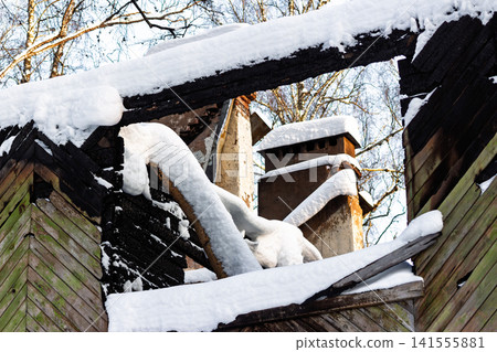 Charred wood structure peeks through heavy winter snowdrifts, framing a view of brick chimneys and stark winter trees against a bright sky Charred wood structure peeks through heavy winter snowdrifts, framing a view of brick chimneys and stark winter trees against a bright sky 141555881