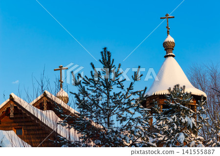Wooden Orthodox church domes capped with fresh snow rise against a bright winter blue sky, framed by evergreen branches in a crisp Russian landscape Wooden Orthodox church domes capped with fresh snow rise against a bright winter blue sky, framed by evergreen branches in a crisp Russian landscape 141555887