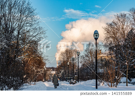 Vibrant winter scene with thick steam plume rising above snow-covered trees under a bright blue sky, single pedestrian walks down a frosty path 141555889