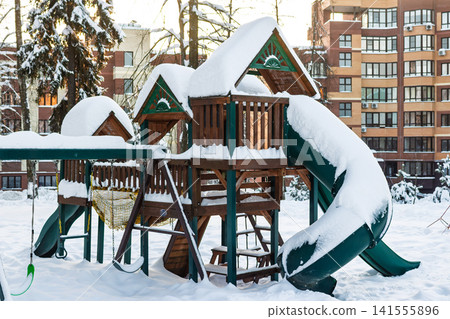 Wooden playground structure buried under thick white snow, deserted winter scene with apartment buildings in the background Wooden playground structure buried under thick white snow, deserted winter scene with apartment buildings in the background 141555896