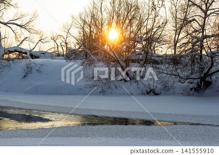 Brilliant winter sunset glowing through bare branches above a partially frozen river, creating a serene, frosty landscape scene Brilliant winter sunset glowing through bare branches above a partially frozen river, creating a serene, frosty landscape scene 141555910