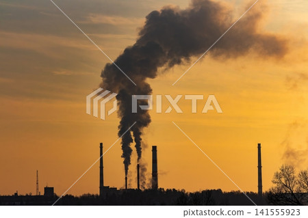 Dark smoke billows from tall industrial smokestacks against a vibrant golden sunset sky, silhouetting the factory skyline and trees below Dark smoke billows from tall industrial smokestacks against a vibrant golden sunset sky, silhouetting the factory skyline and trees below 141555923