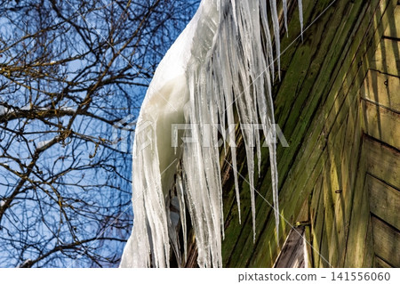 Huge, sharp icicles hang menacingly from an old timber house eave, a winter hazard. Freezing danger, brittle beauty against blue sky Huge, sharp icicles hang menacingly from an old timber house eave, a winter hazard. Freezing danger, brittle beauty against blue sky 141556060