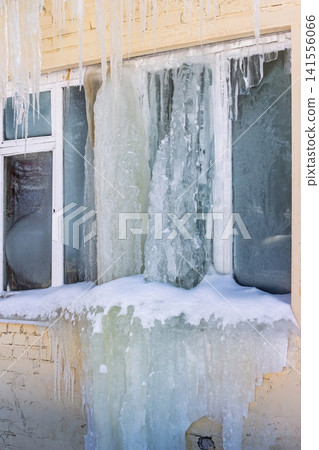 Massive ice formations encase a window and wall, with heavy icicles dangling from the roofline. A striking winter spectacle showcasing freezing temps Massive ice formations encase a window and wall, with heavy icicles dangling from the roofline. A striking winter spectacle showcasing freezing temps 141556066