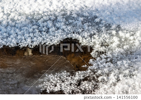 Intricate frost patterns edge a dark, open stream section in a close-up winter scene, showing icy textures and brown water contrast Intricate frost patterns edge a dark, open stream section in a close-up winter scene, showing icy textures and brown water contrast 141556100