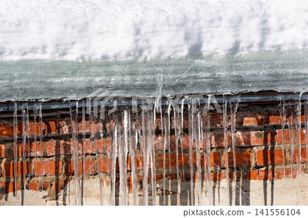 Frozen daggers of ice drip above a rustic brick wall, bathed in bright winter sun. Danger of melting snow and ice Frozen daggers of ice drip above a rustic brick wall, bathed in bright winter sun. Danger of melting snow and ice 141556104