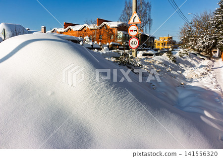 Pristine white snow mounds heap along the sunlit highway shoulder, with urban rooftops peeking under a vibrant winter sky Pristine white snow mounds heap along the sunlit highway shoulder, with urban rooftops peeking under a vibrant winter sky 141556320