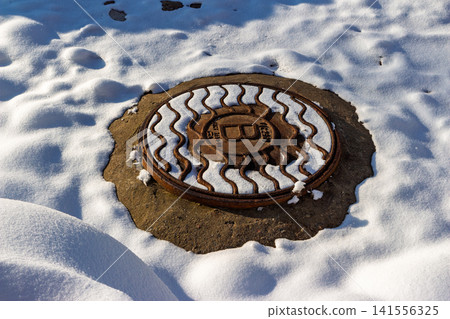 Rusty old cast iron manhole cover partially buried in bright white snow during winter sunshine, Russia Rusty old cast iron manhole cover partially buried in bright white snow during winter sunshine, Russia 141556325