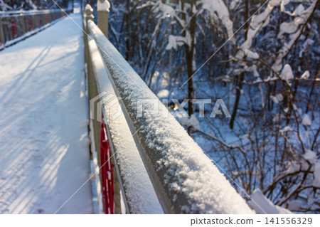 Sparkling snow blankets a pedestrian bridge railing, leading along a frosty path. Winter wonderland scene with soft light and bare trees Sparkling snow blankets a pedestrian bridge railing, leading along a frosty path. Winter wonderland scene with soft light and bare trees 141556329