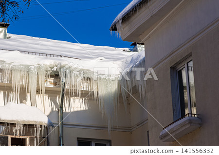 Dramatic, huge icicles gleam on a thick blanket of rooftop snow against a vibrant winter sky, showcasing nature's cold beauty and potential danger Dramatic, huge icicles gleam on a thick blanket of rooftop snow against a vibrant winter sky, showcasing nature's cold beauty and potential danger 141556332