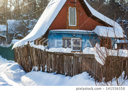 Rustic dacha house under a heavy blanket of fresh snow, sunlight illuminating the charming winter scene. A wooden fence in the foreground adds to the cozy, old-world vibe Rustic dacha house under a heavy blanket of fresh snow, sunlight illuminating the charming winter scene. A wooden fence in the foreground adds to the cozy, old-world vibe 141556350