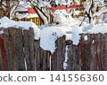 Weathered wooden fence adorned with thick, sparkling snow under a bright winter sun, capturing a cozy, rustic village scene 141556360