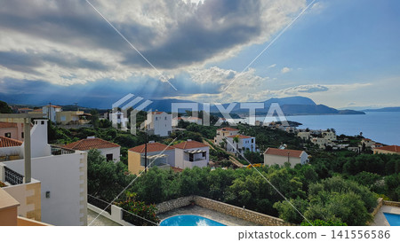 Scenic view of coastal Greek village with white houses and sunbeams breaking through dramatic clouds over sea in Crete for travel destination stock background Scenic view of coastal Greek village with white houses and sunbeams breaking through dramatic clouds over sea in Crete for travel destination stock background 141556586