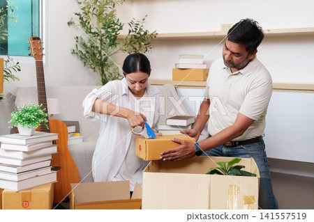 Indian couple packing cardboard box with tape dispenser for moving house Indian couple packing cardboard box with tape dispenser for moving house 141557519