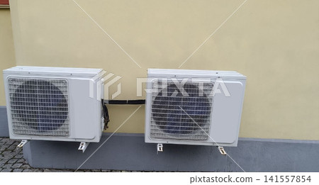Two white outdoor air conditioning units mounted on beige wall with gray base. Two white outdoor air conditioning units mounted on beige wall with gray base. 141557854