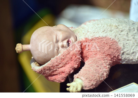 Very old plastic baby doll with a crying expression wearing a worn and stained sleeper, displayed for sale at an outdoor flea market Very old plastic baby doll with a crying expression wearing a worn and stained sleeper, displayed for sale at an outdoor flea market 141558069