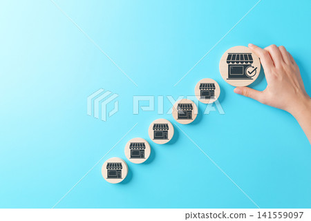 Business Growth and Franchise Expansion Concept. Hand placing large wooden block with store icon and check mark among smaller store icons, symbolizing franchise development, retail expansion, strategy Business Growth and Franchise Expansion Concept. Hand placing large wooden block with store icon and check mark among smaller store icons, symbolizing franchise development, retail expansion, strategy 141559097