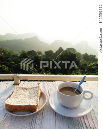 A cup of hot coffee with toast bread in white dish on white wooden counter bar is breakfast in resort on the mountain in Thailand A cup of hot coffee with toast bread in white dish on white wooden counter bar is breakfast in resort on the mountain in Thailand 141559212