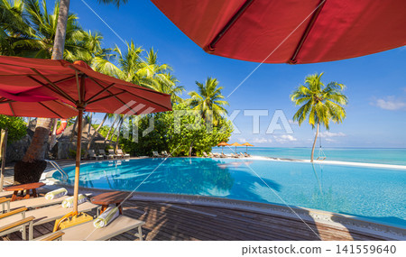 Luxurious tropical poolside with red umbrellas and loungers under palm trees beside turquoise lagoon. Perfect summer vacation travel resort paradise. Luxury getaway relax leisure scenic Luxurious tropical poolside with red umbrellas and loungers under palm trees beside turquoise lagoon. Perfect summer vacation travel resort paradise. Luxury getaway relax leisure scenic 141559640