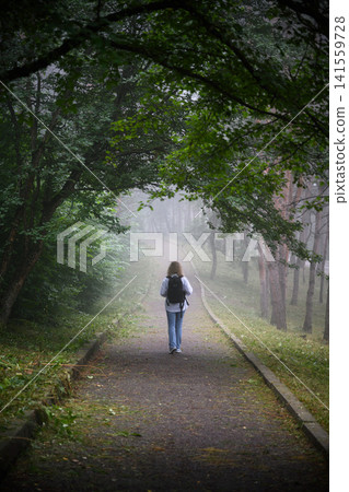 Woman with a black backpack walking away on a foggy forest path. Vertical view of a female hiking on misty park trail in the morning. Journey and adventure concept Woman with a black backpack walking away on a foggy forest path. Vertical view of a female hiking on misty park trail in the morning. Journey and adventure concept 141559728