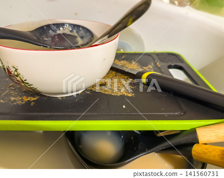 Dirty dishes and utensils stacked on cutting board in kitchen sink 141560731