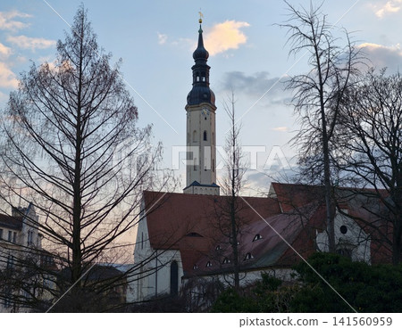 Ancient church tower with bare trees and pastel sunset illuminating tranquil village scene Ancient church tower with bare trees and pastel sunset illuminating tranquil village scene 141560959