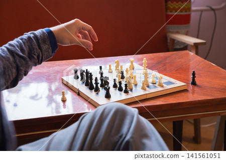 Man playing chess on wooden table. Gamer moving piece on chessboard. Logical thinking and strategy game. Indoor leisure activity and intellectual hobby for entertainment in home environment. Man playing chess on wooden table. Gamer moving piece on chessboard. Logical thinking and strategy game. Indoor leisure activity and intellectual hobby for entertainment in home environment. 141561051