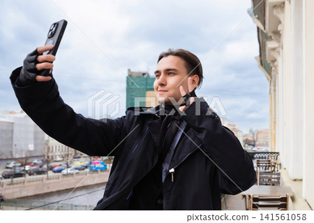 Man taking selfie outdoors on the balcony. Young fashionable guy holding smartphone for photography. Stylish person wearing black coat and gloves posing for digital picture concept. Man taking selfie outdoors on the balcony. Young fashionable guy holding smartphone for photography. Stylish person wearing black coat and gloves posing for digital picture concept. 141561058