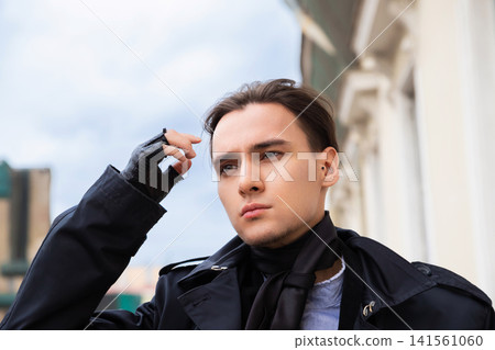 Man in black trench coat wearing fingerless gloves looking away. Portrait of stylish young man outdoors. Fashion concept, urban street lifestyle, trendy male clothing and accessories. Man in black trench coat wearing fingerless gloves looking away. Portrait of stylish young man outdoors. Fashion concept, urban street lifestyle, trendy male clothing and accessories. 141561060