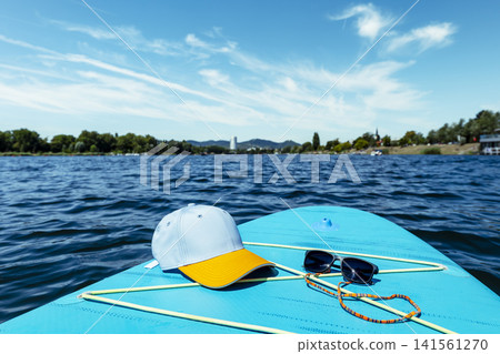 Baseball cap and sunglasses on a turquoise stand up paddleboard, illustrating summer water sport and outdoor leisure. 141561270