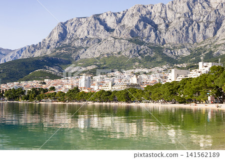 Makarska town and its vibrant beach line the Adriatic Sea, reflecting in the clear waters with the dramatic Biokovo Mountains creating a stunning backdrop Makarska town and its vibrant beach line the Adriatic Sea, reflecting in the clear waters with the dramatic Biokovo Mountains creating a stunning backdrop 141562189