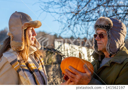 Man and woman holding pumpkin. Couple wearing fur hat outdoor. Autumn harvest celebration concept. Seasonal vegetables for thanksgiving holiday and healthy food lifestyle. Man and woman holding pumpkin. Couple wearing fur hat outdoor. Autumn harvest celebration concept. Seasonal vegetables for thanksgiving holiday and healthy food lifestyle. 141562197