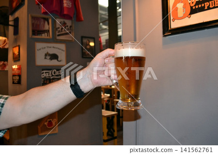 Hand holding a beer mug with frothy head in a cozy pub interior for bar menu or social media banner and beverage promotion Hand holding a beer mug with frothy head in a cozy pub interior for bar menu or social media banner and beverage promotion 141562761