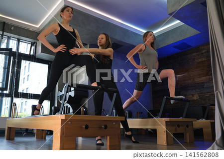 A trainer assists a woman balancing on a Pilates reformer while another client repeats the pose nearby. The scene reflects boutique fitness service, personal guidance, mobility work, and quality care. A trainer assists a woman balancing on a Pilates reformer while another client repeats the pose nearby. The scene reflects boutique fitness service, personal guidance, mobility work, and quality care. 141562808