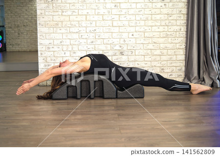A fit woman stretches deeply over a Pilates spine corrector beside a white brick wall. The scene connects flexibility, back health, recovery routines, emotional calm, and self care. A fit woman stretches deeply over a Pilates spine corrector beside a white brick wall. The scene connects flexibility, back health, recovery routines, emotional calm, and self care. 141562809
