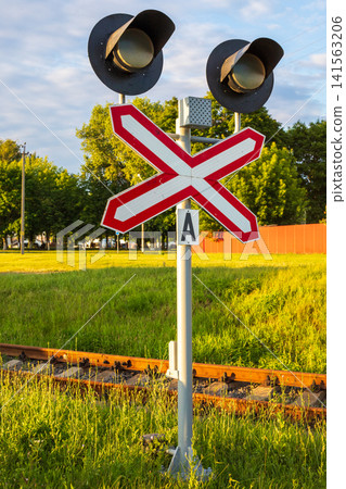 Railway crossing signal post with crossbuck sign and dual traffic lights, railroad track background in summer evening light Railway crossing signal post with crossbuck sign and dual traffic lights, railroad track background in summer evening light 141563206