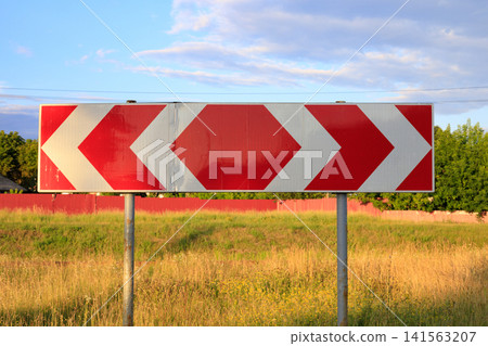 Red and white chevron directional road sign on posts with field and blue sky background, hazard warning chevron pointer Red and white chevron directional road sign on posts with field and blue sky background, hazard warning chevron pointer 141563207