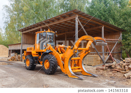 Orange forestry loader with a claw attachment parked at a sawmill near a wooden shed Orange forestry loader with a claw attachment parked at a sawmill near a wooden shed 141563318