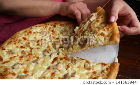 A woman's hands grasp a delicious triangular slice of round Hawaiian pizza with a fluffy edge. The filling consists of juicy chicken breast, diced pineapple, and baked, melted mozzarella cheese. A woman's hands grasp a delicious triangular slice of round Hawaiian pizza with a fluffy edge. The filling consists of juicy chicken breast, diced pineapple, and baked, melted mozzarella cheese. 141563944