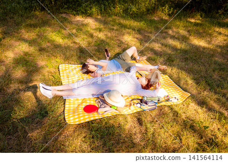 Middle-aged mother and her adult daughter relaxing on a yellow plaid in a summer park, enjoying nature and bonding. Concept of family vacation and mental support. Middle-aged mother and her adult daughter relaxing on a yellow plaid in a summer park, enjoying nature and bonding. Concept of family vacation and mental support. 141564114