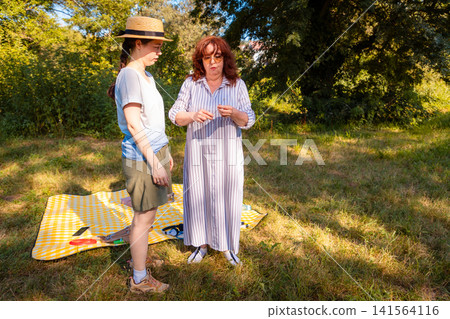 Caucasian mother and adult daughter share a relaxing summer picnic in the park enjoying conversation, snacks, and quality family time in nature. Wide shot Caucasian mother and adult daughter share a relaxing summer picnic in the park enjoying conversation, snacks, and quality family time in nature. Wide shot 141564116