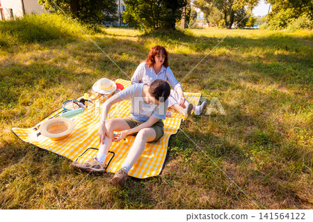 Young woman and her mother resting outdoors and scratching skin due to mosquito attacks during picnic time in public summer park. Wide shot Young woman and her mother resting outdoors and scratching skin due to mosquito attacks during picnic time in public summer park. Wide shot 141564122