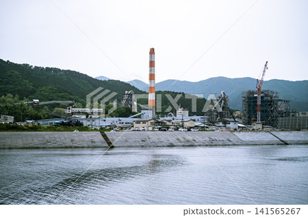 Under a cloudy sky, a close-up shot with a 45mm lens captures the scenery of the seawall and mountain range, with red and white chimneys and factory plants towering over the landscape. Under a cloudy sky, a close-up shot with a 45mm lens captures the scenery of the seawall and mountain range, with red and white chimneys and factory plants towering over the landscape. 141565267