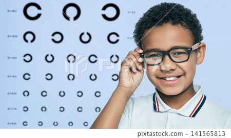 Smiling African American boy touching modern glasses frame during vision test. Pediatric ophthalmology diagnostic consultation and myopia correction. Smiling African American boy touching modern glasses frame during vision test. Pediatric ophthalmology diagnostic consultation and myopia correction. 141565813