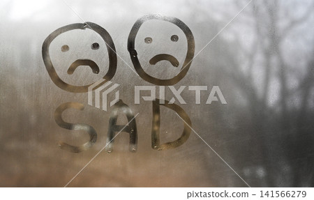 Sad word appears on fogged window glass with two unhappy faces. Sad word appears on fogged window glass with two unhappy faces. 141566279