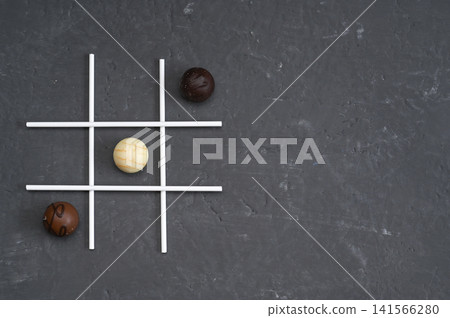 Chocolate candies arranged as tic tac toe game on dark stone background with white grid lines. Chocolate balls act as players pieces creating playful strategy mood. concept for fun dessert, game idea Chocolate candies arranged as tic tac toe game on dark stone background with white grid lines. Chocolate balls act as players pieces creating playful strategy mood. concept for fun dessert, game idea 141566280
