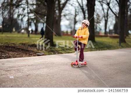 Happy elementary age girl riding scooter in park Happy elementary age girl riding scooter in park 141566759